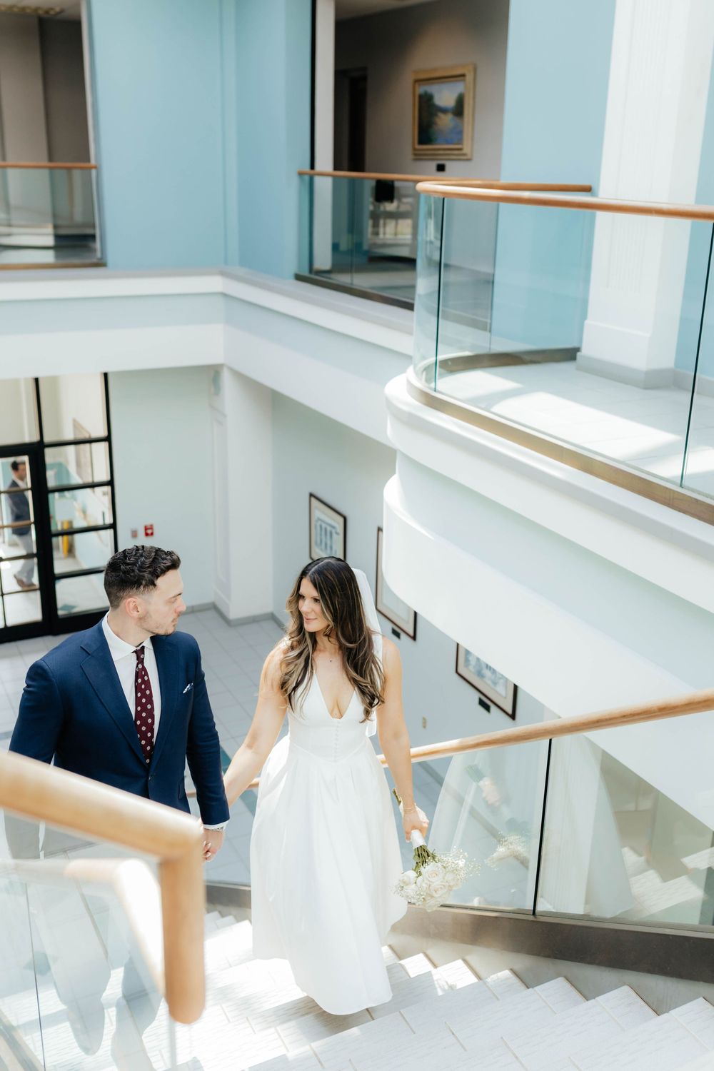 Couple in formal wedding attire sharing a romantic moment in a modern stairwell with glass railings.