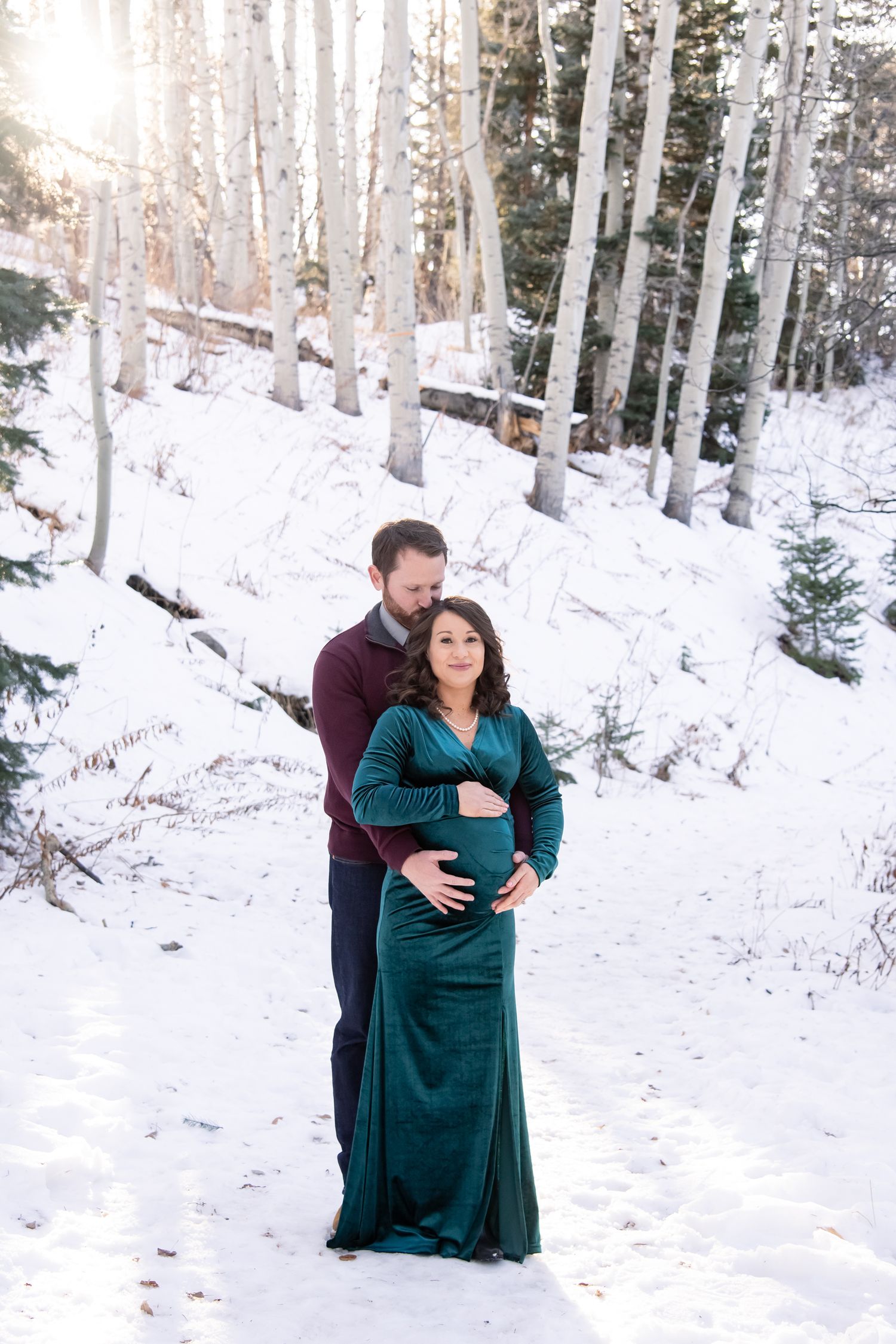 A couple in a winter setting shares an intimate moment as they cradle a pregnant belly in an emerald green dress for Aspen CO maternity