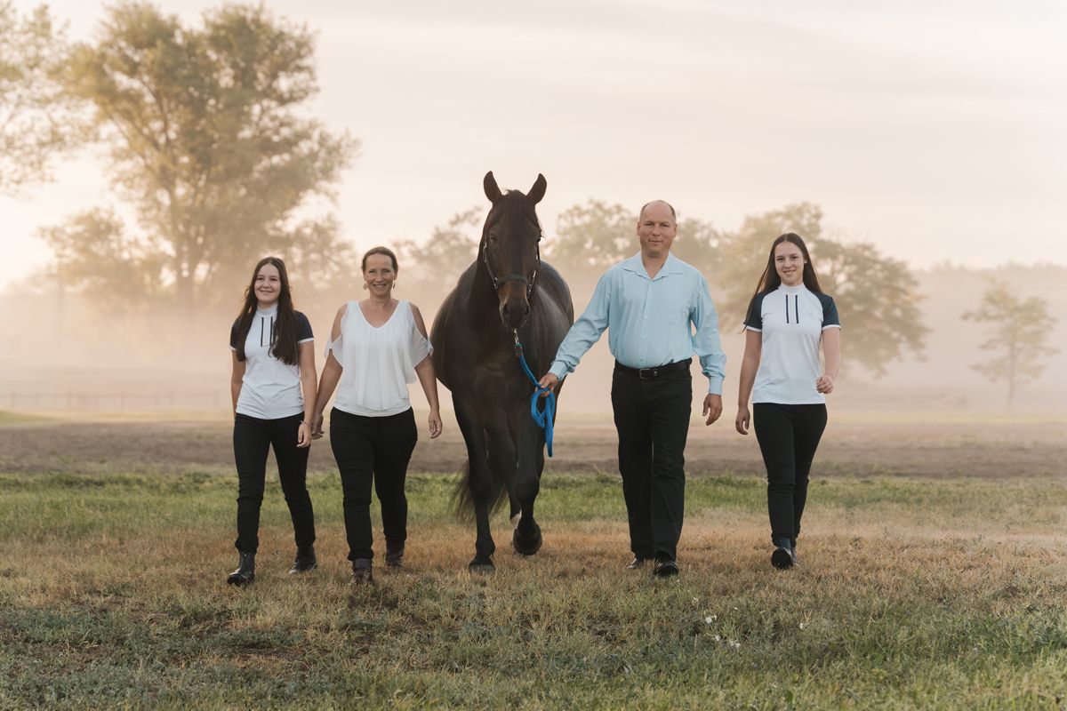 A family of 4 walking together through a misty field at sunrise, holding hands in a line formation with their horse during a photoshoot