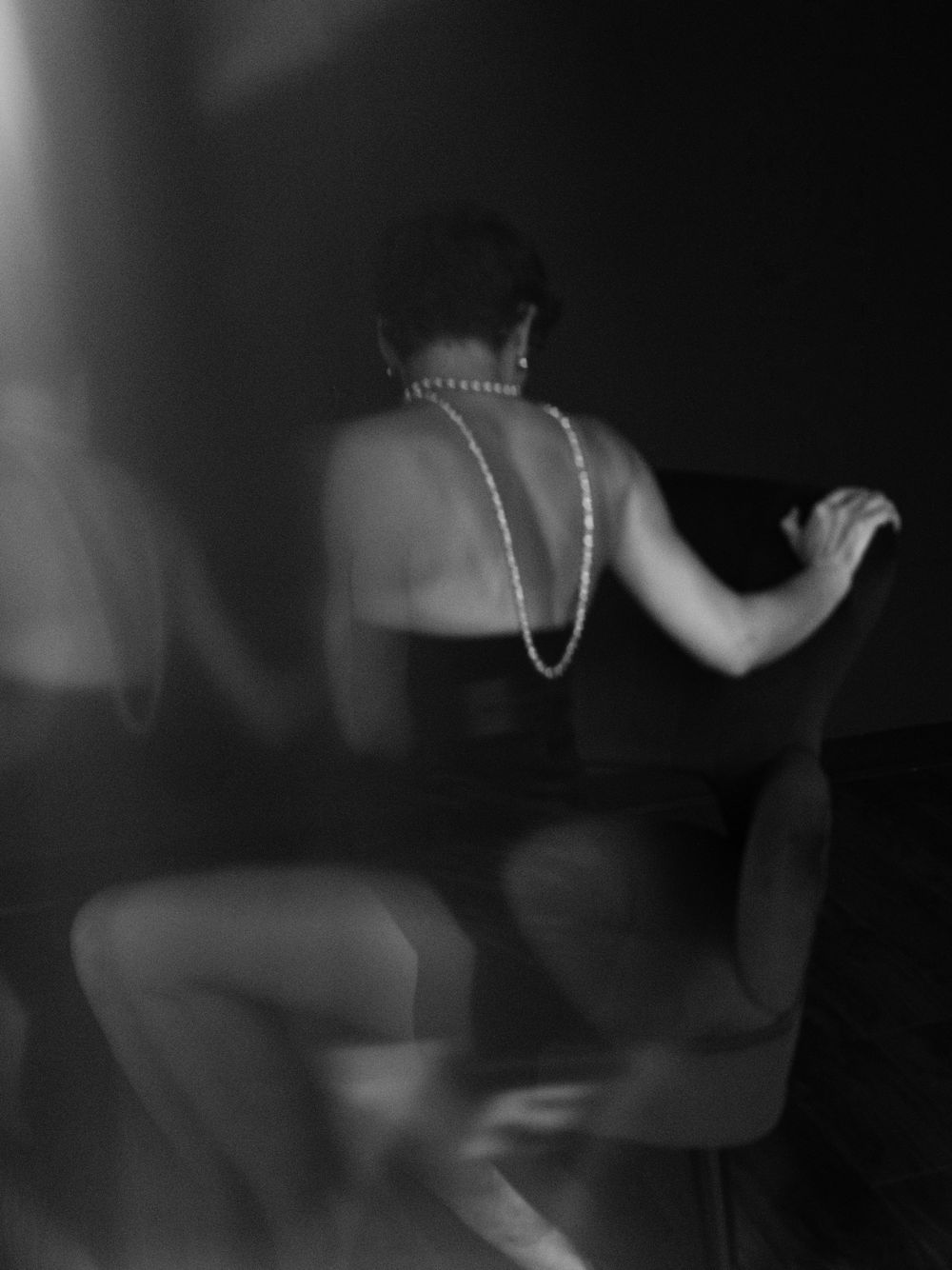 A black and white artistic photograph showing a figure from behind wearing a pearl necklace in a dark, moody setting.