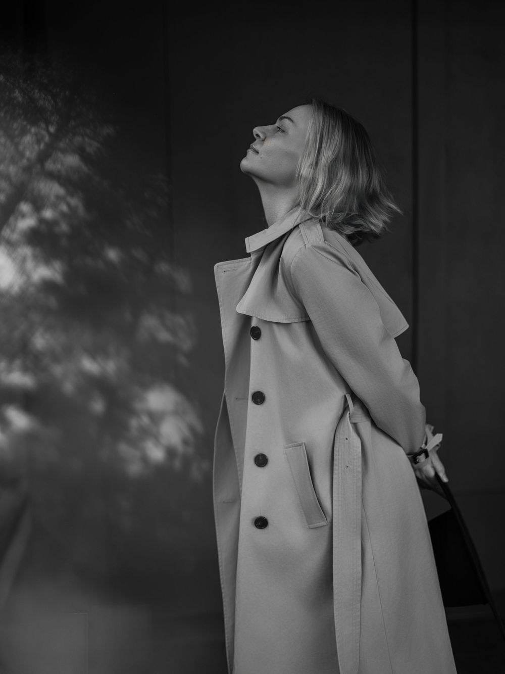 Black and white fashion portrait of a person in a light-colored trench coat looking up against a shadowy background.