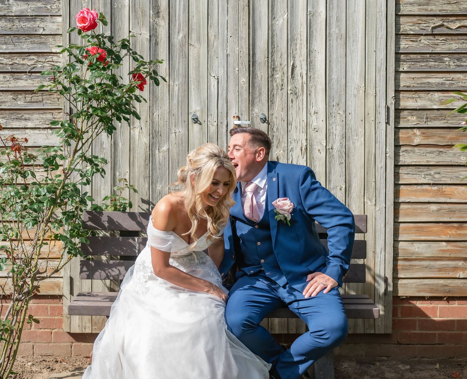 A couple in wedding attire shares a romantic moment on a rustic wooden bench surrounded by roses.