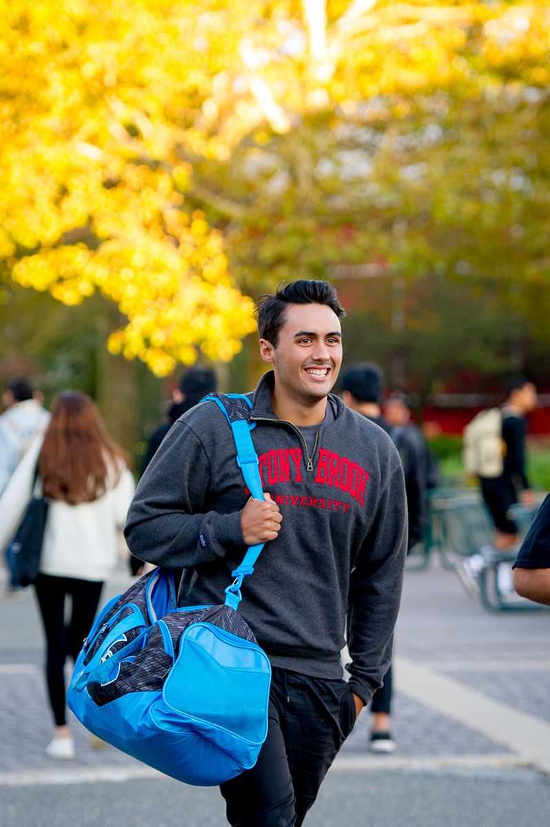Student wearing gray sweatshirt and carrying bright blue duffle bag smiles while walking across campus in autumn.