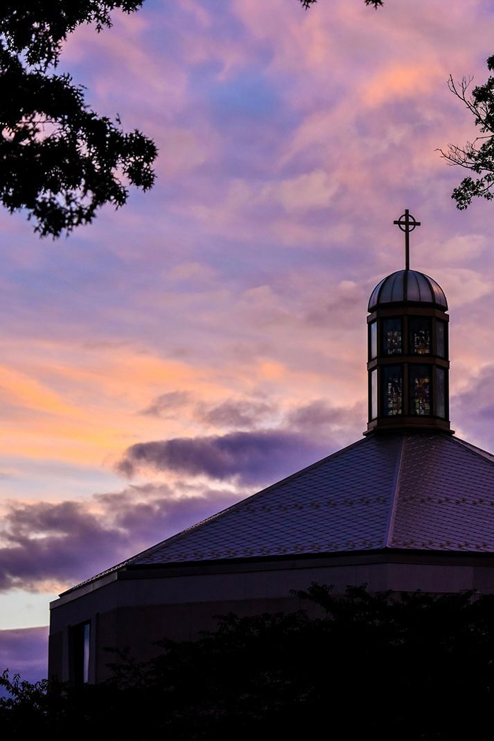 Church steeple silhouetted against a dramatic purple and orange sunset sky with tree branches framing the scene.