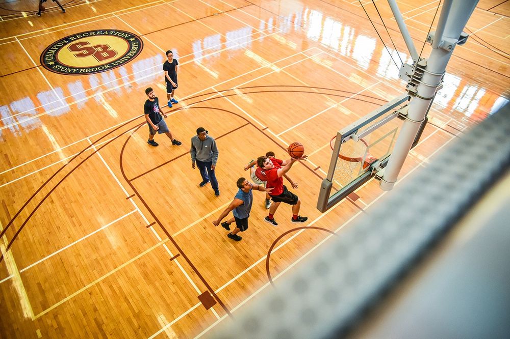 Basketball players compete on a shiny hardwood court viewed from above through protective netting.