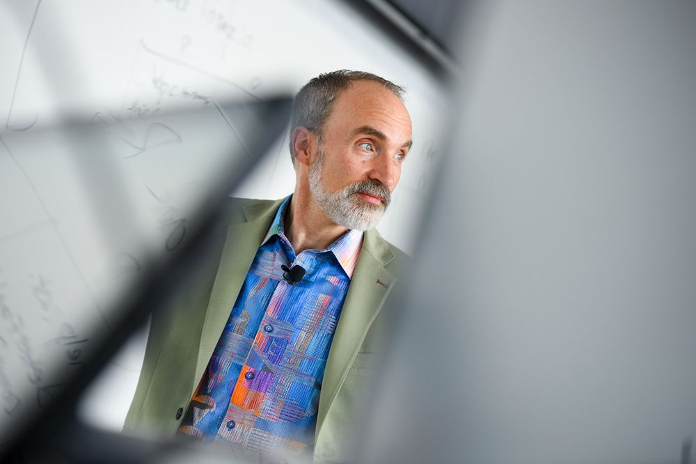 A person in a beige blazer and blue shirt viewed through an angular frame or window.