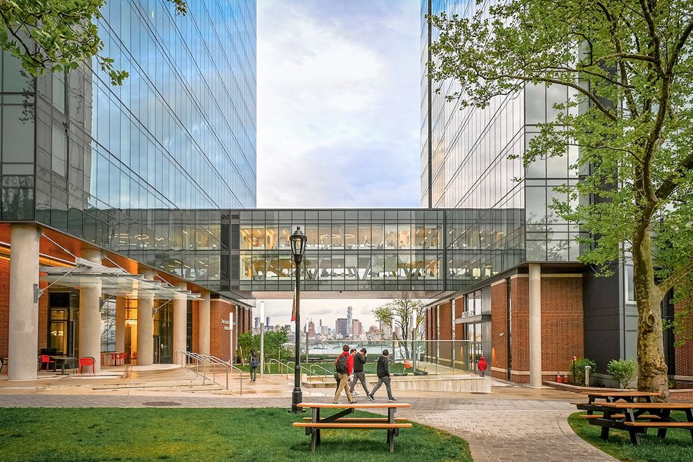 Modern office building exterior features glass walls and wooden benches surrounded by green trees in an urban setting.