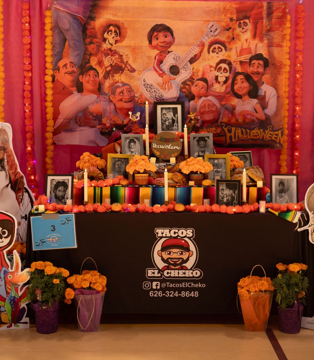 Colorful Day of the Dead altar display with marigolds, photos, and decorations on a black-draped table against an orange backdrop.