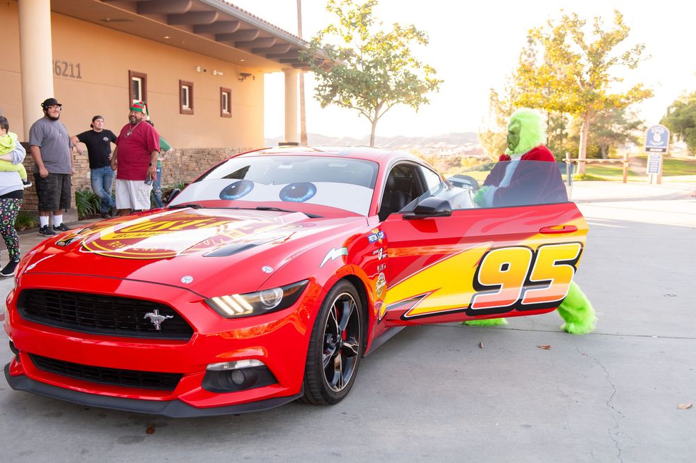 A row of red Ford Mustang sports cars parked in a line at an outdoor event with spectators visible in background.