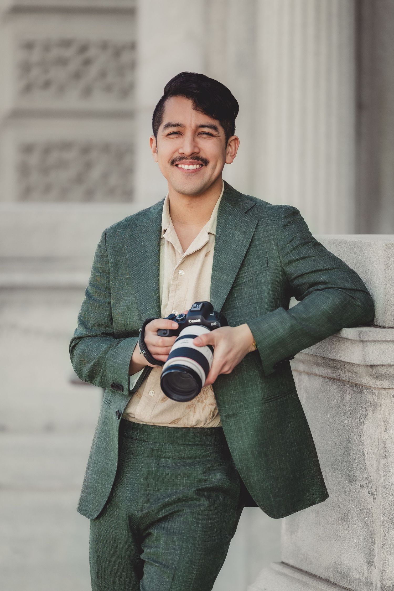 Professional photographer in green suit holding camera and posing against white stone columns.