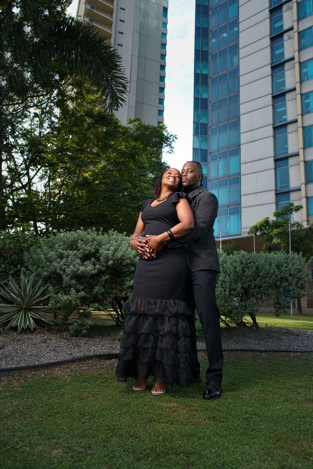 A romantic couple poses together in front of a modern high-rise building with palm trees and landscaping.