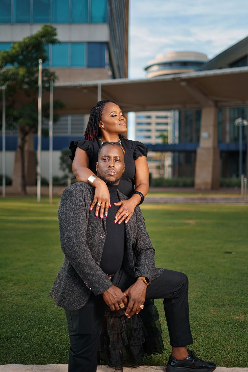 Two people in black outfits pose together on grass with modern buildings in the background during golden hour.