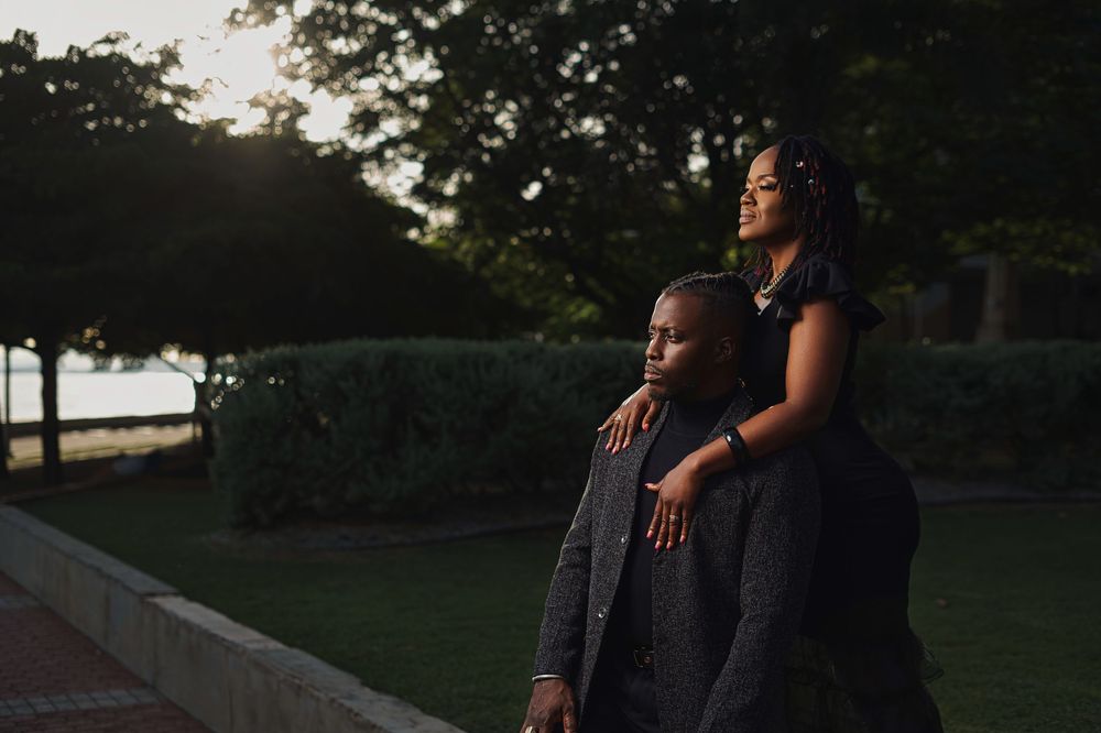A couple embraces while standing outdoors at sunset with trees and hedges in the background.