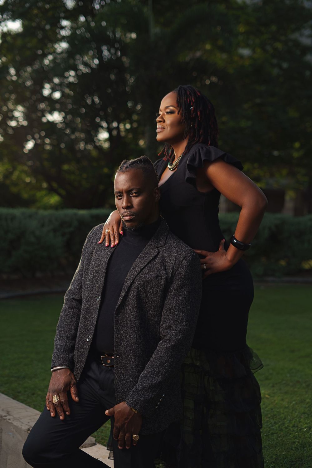 A stylish couple in formal attire poses together against a green outdoor background.