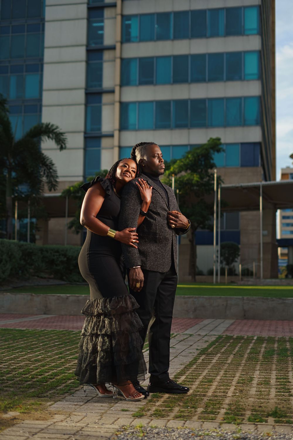 A couple shares an intimate moment while sitting together on patterned pavement outside a modern office building.