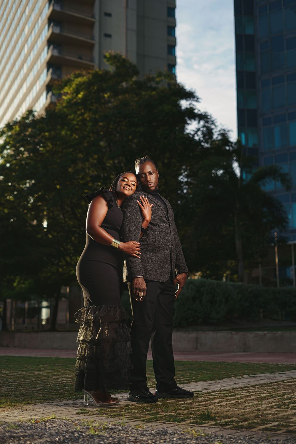 A couple in elegant black attire poses for a romantic portrait against city buildings at dusk.