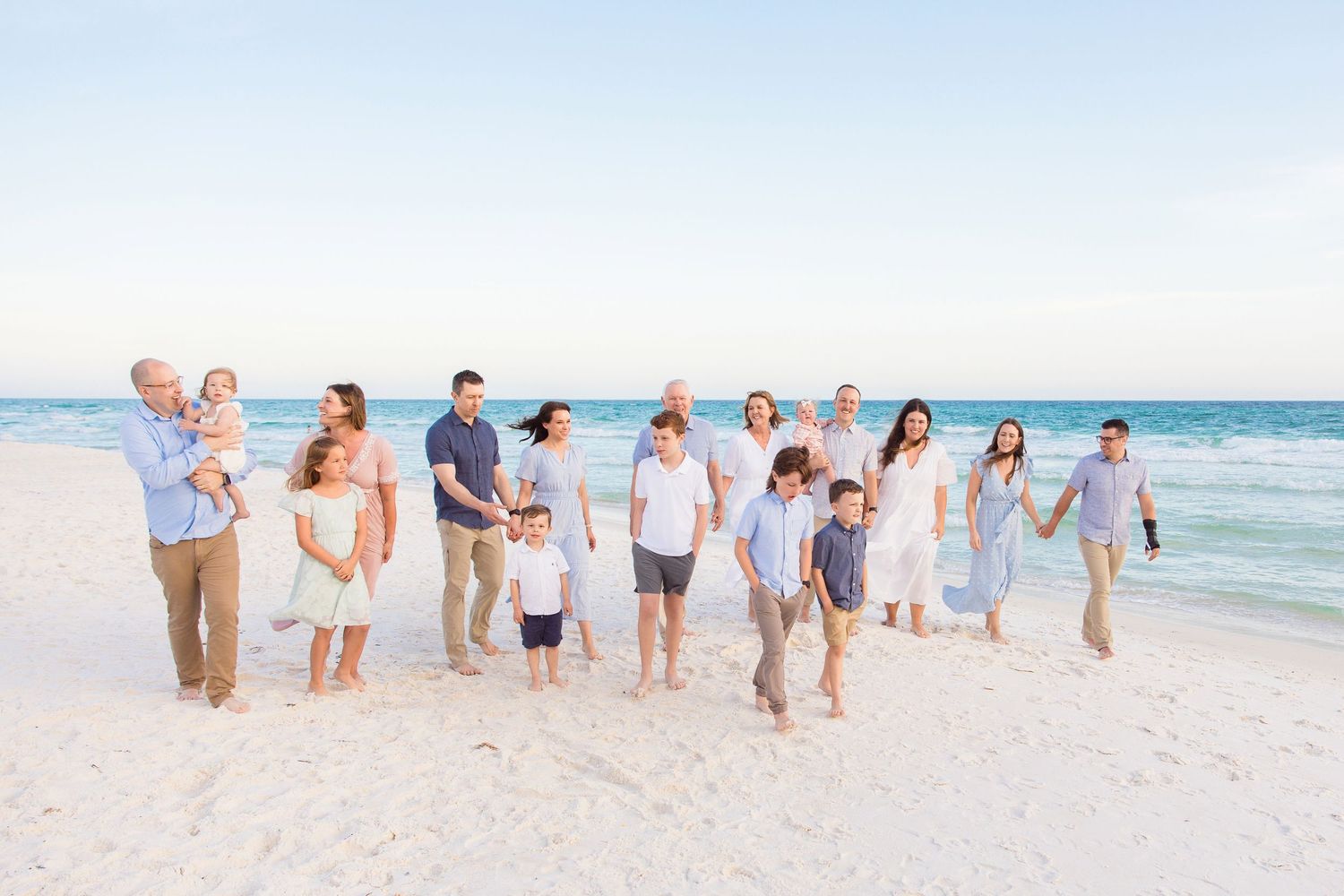 A group of people in light colored clothing walk together along a sunny beach with ocean waves in the background.