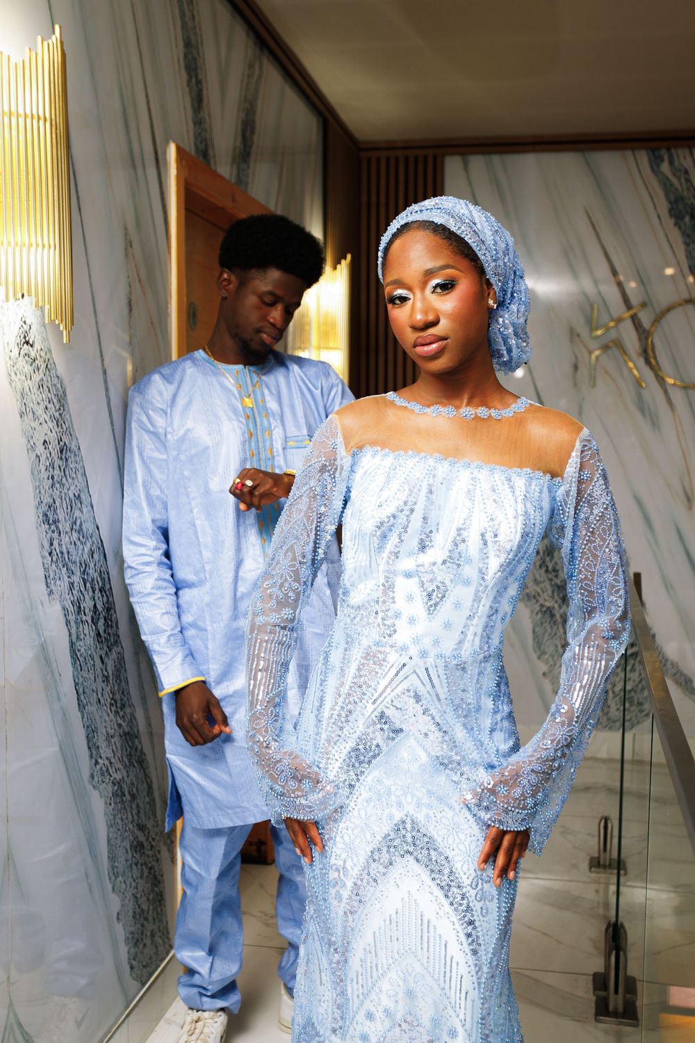 A couple in matching light blue traditional African attire poses together in an elegant indoor setting.