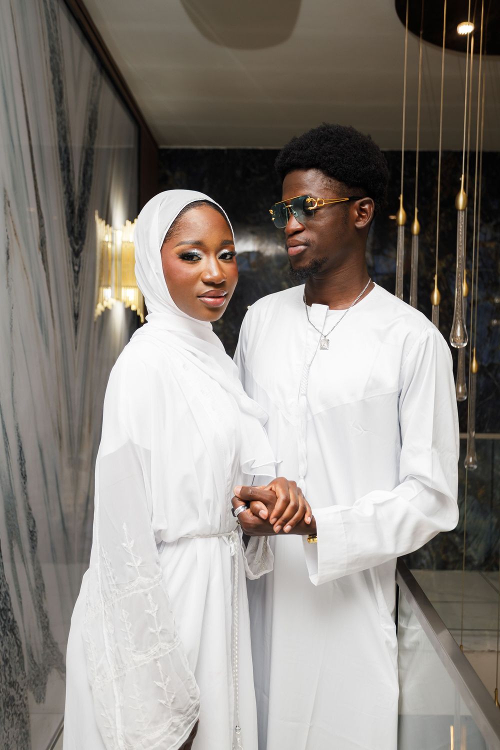 A couple in matching white outfits share intimate moments in an elegant indoor setting with curtained windows.