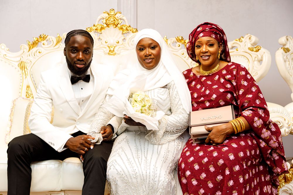 Three people in formal attire sit together at a wedding celebration with elegant white decor in the background.