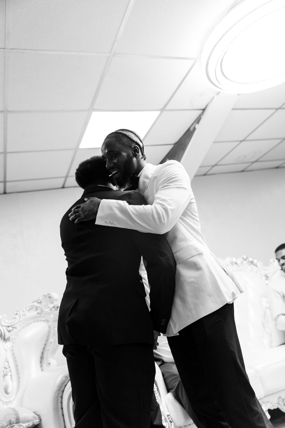 Two figures in black and white attire embrace in an emotional moment inside a bright white room.