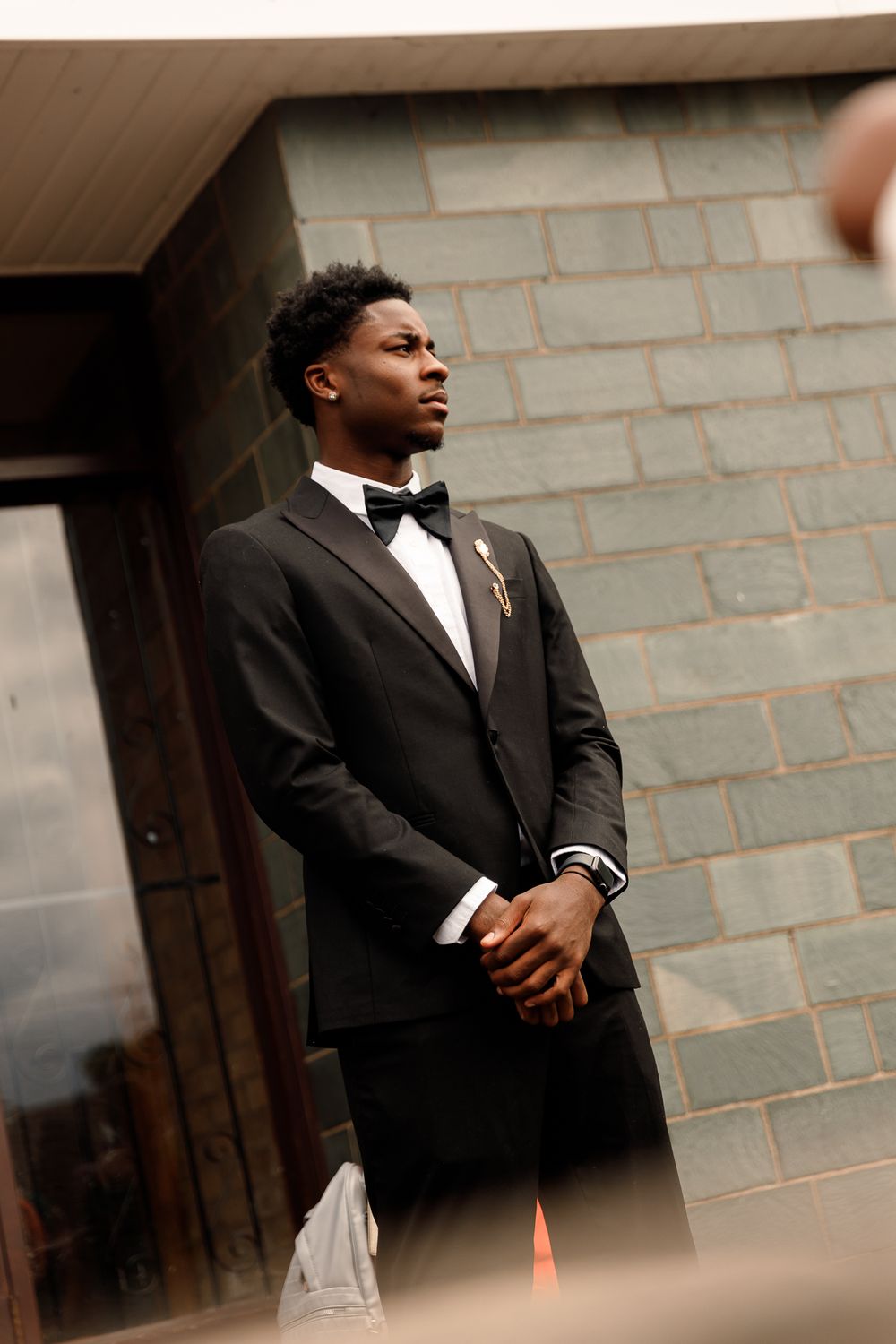 A young man in a formal black suit poses confidently against a brick wall in a series of professional portraits.