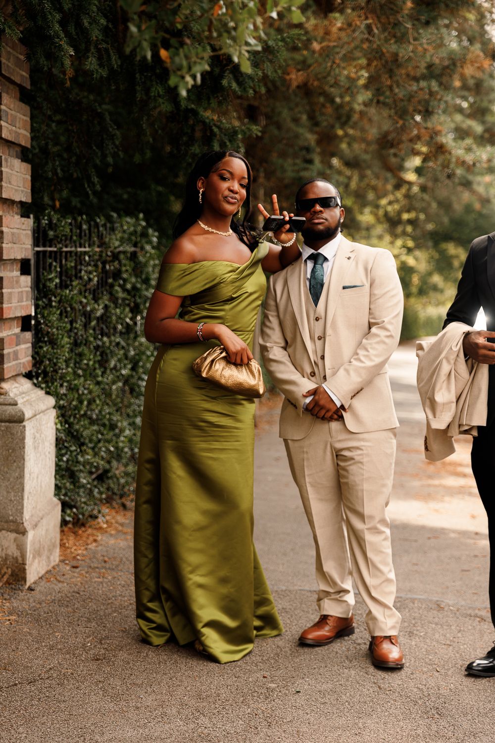 A couple poses together outdoors, with one wearing an olive green dress and the other in a beige suit.