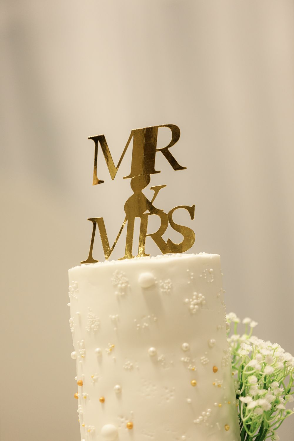 White wedding cake decorated with gold 'MR & MRS' topper and delicate floral accents.