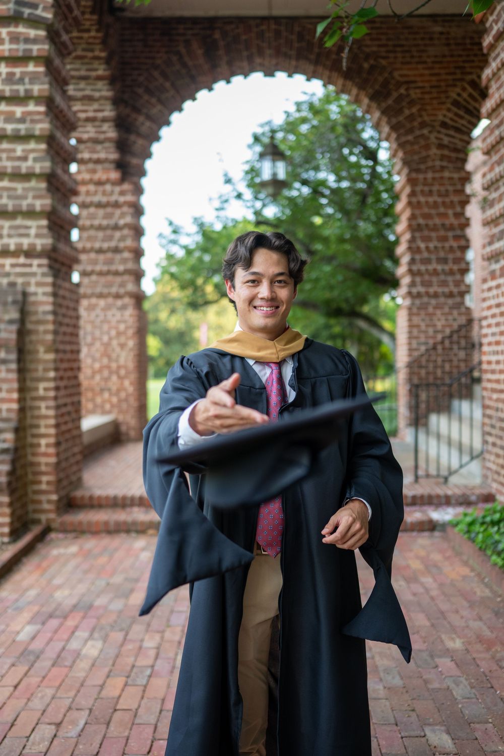 Graduate in academic regalia poses with a warm smile under a brick archway on a university campus.