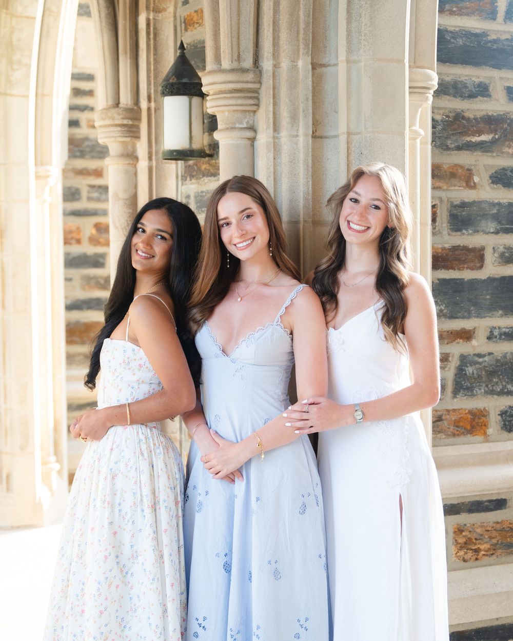 Three young women in elegant pastel dresses posing together under stone archways at a formal event.
