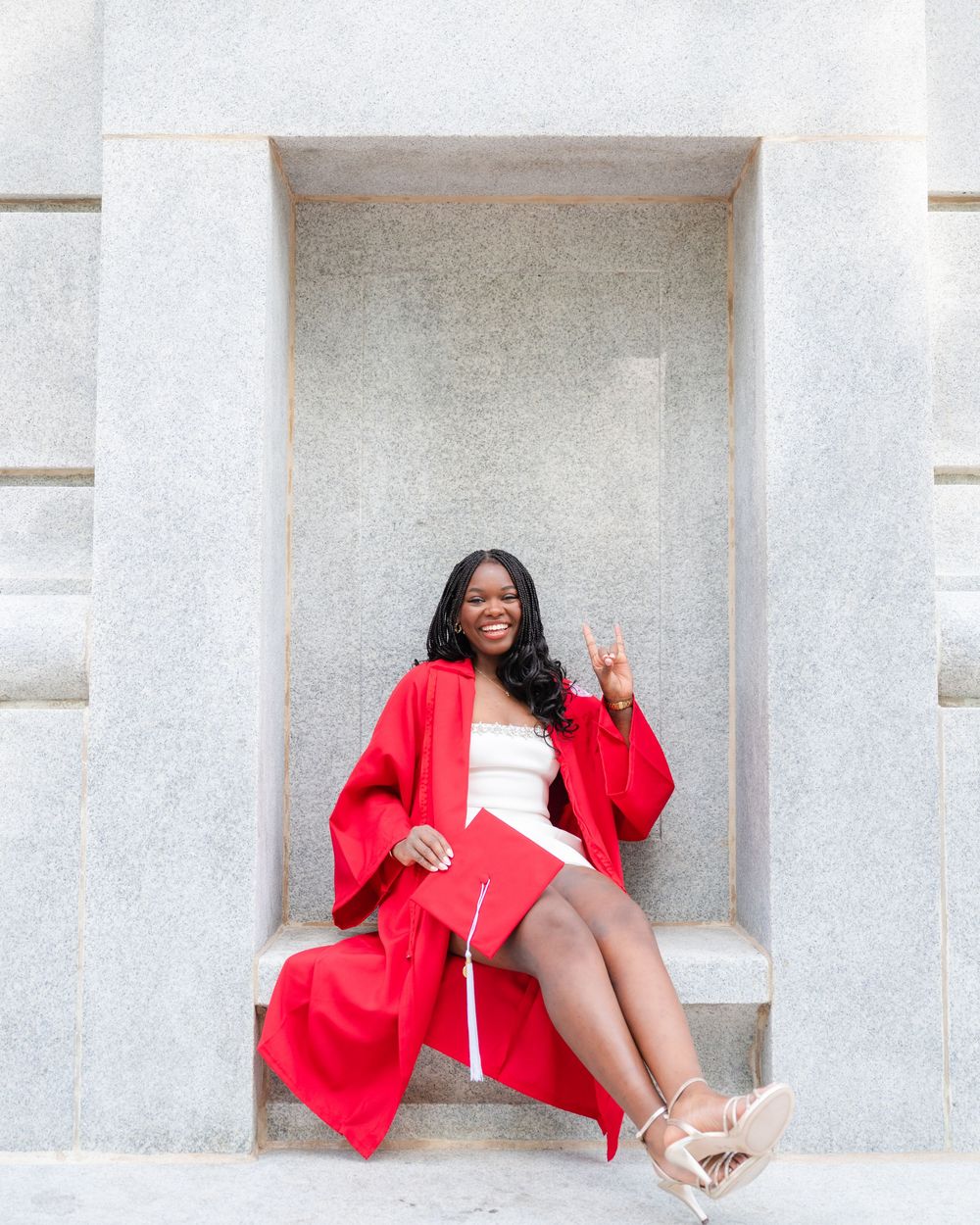 Young woman in red graduation gown and cap smiles while seated in a stone alcove, celebrating her achievement.