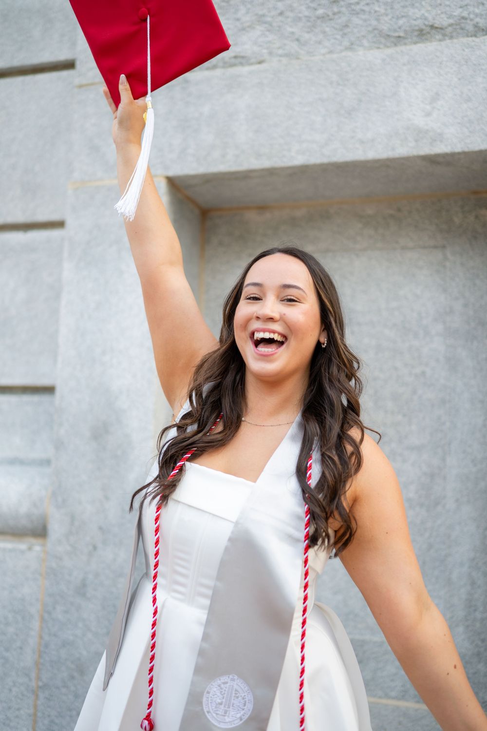 Graduate in white cap and gown raises diploma excitedly during graduation ceremony celebration.