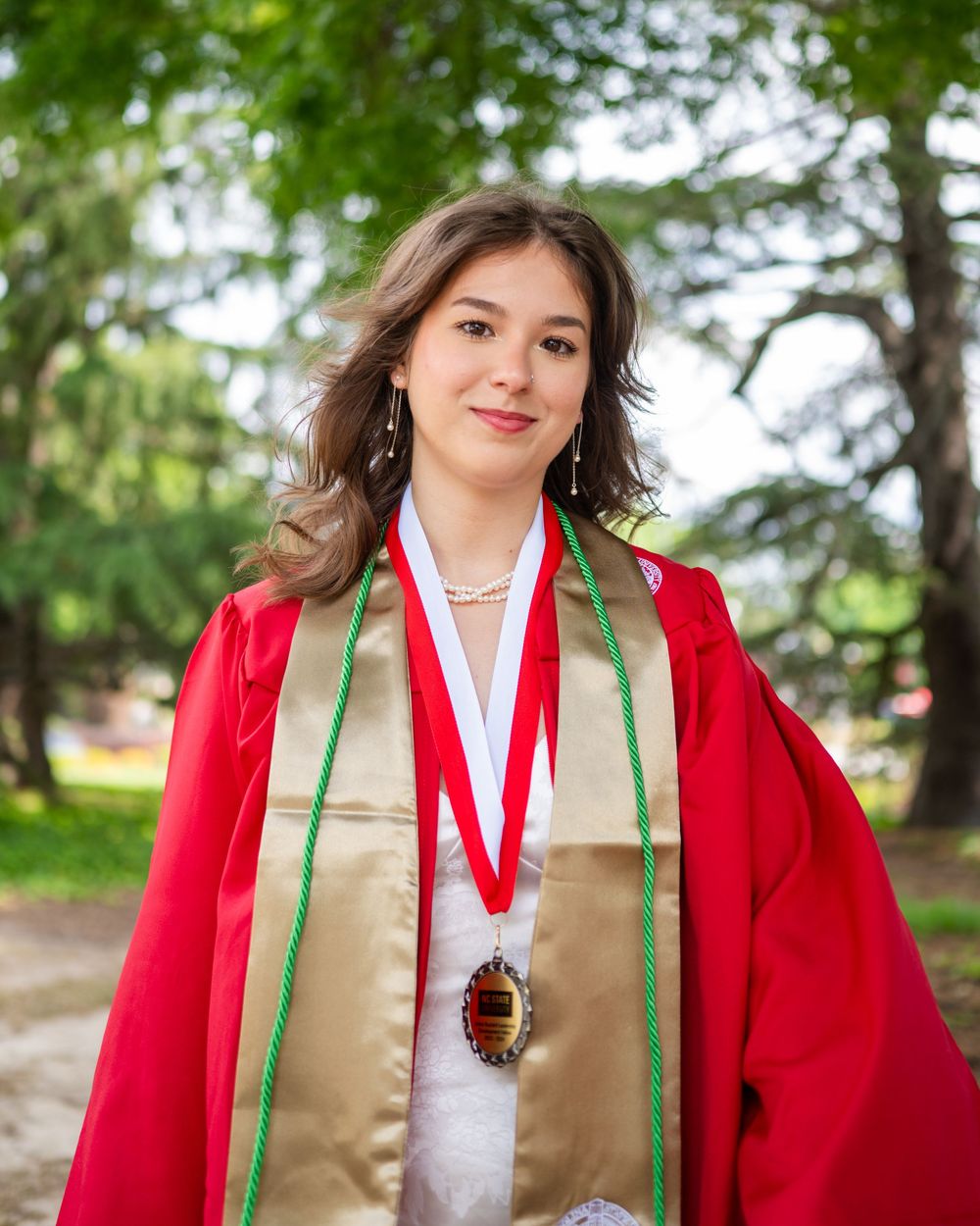 Graduate wearing red academic regalia and gold stole poses outdoors with trees in background during graduation ceremony.