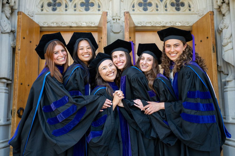 Group of graduates in black caps and gowns with blue stoles pose together during commencement ceremony.