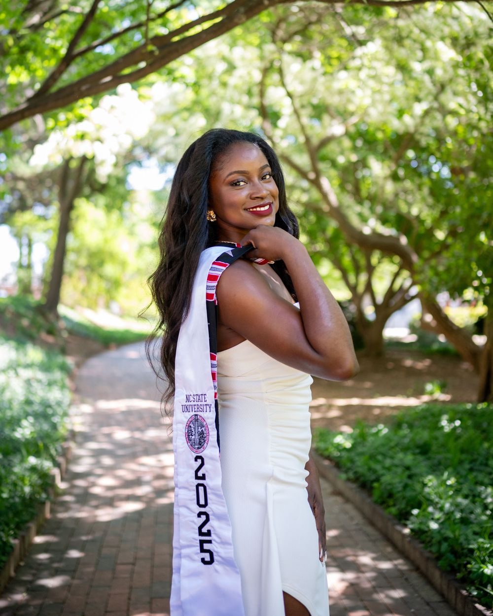 A person in a white dress poses joyfully in a lush garden setting with dappled sunlight filtering through trees.