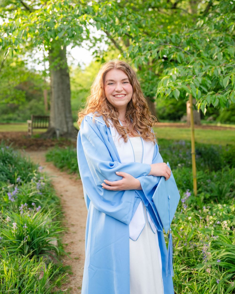 Smiling young woman in light blue graduation gown standing on a garden path surrounded by lush green trees and flowers.