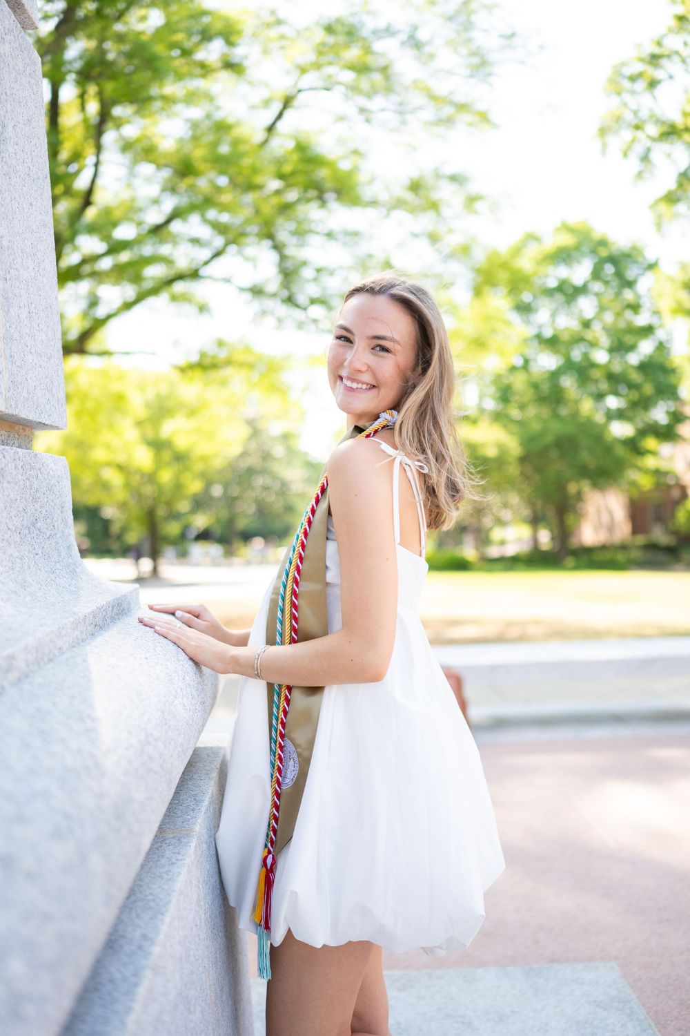 A person in a white summer dress poses outdoors near stone columns on a sunny day with trees in the background.