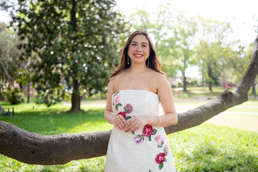 A bride in a white wedding dress poses outdoors in a lush garden setting with blooming trees and natural sunlight.