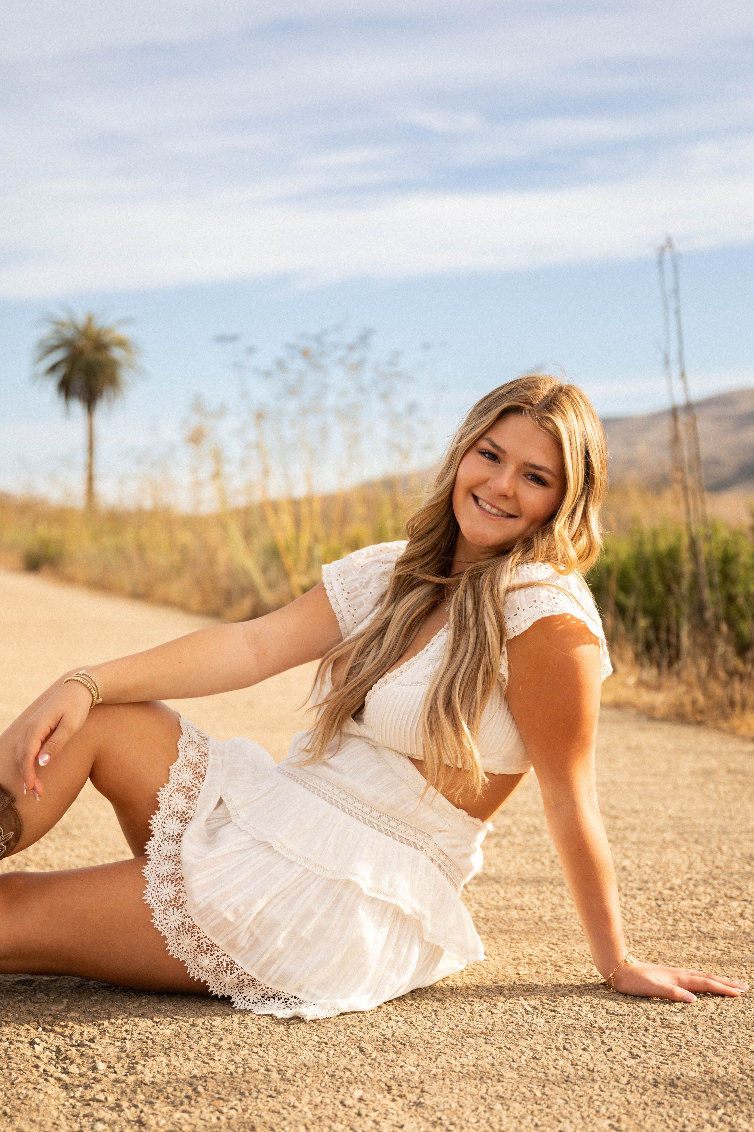A woman in white lace dress relaxes on a dirt path with desert mountains and palm tree backdrop.