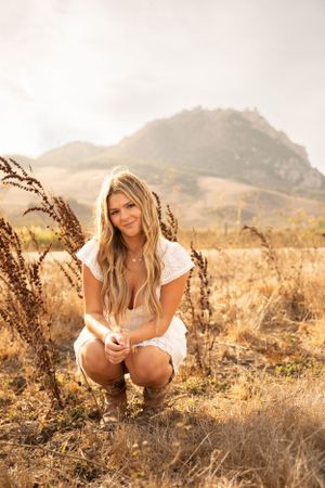 A woman in white crouches among tall grasses with mountains in background during sunset.