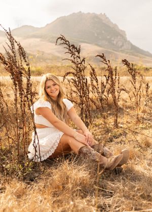 A woman in white sits among wild grasses with mountain backdrop during golden hour.