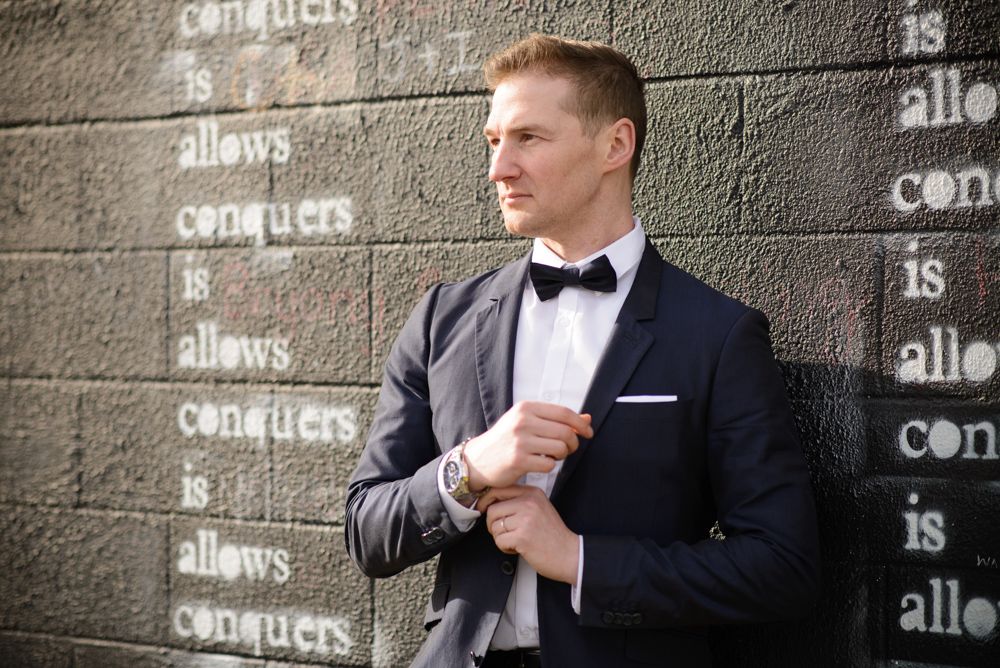 A person in a classic black tuxedo and bow tie adjusts their cuff while standing against a textured concrete wall.
