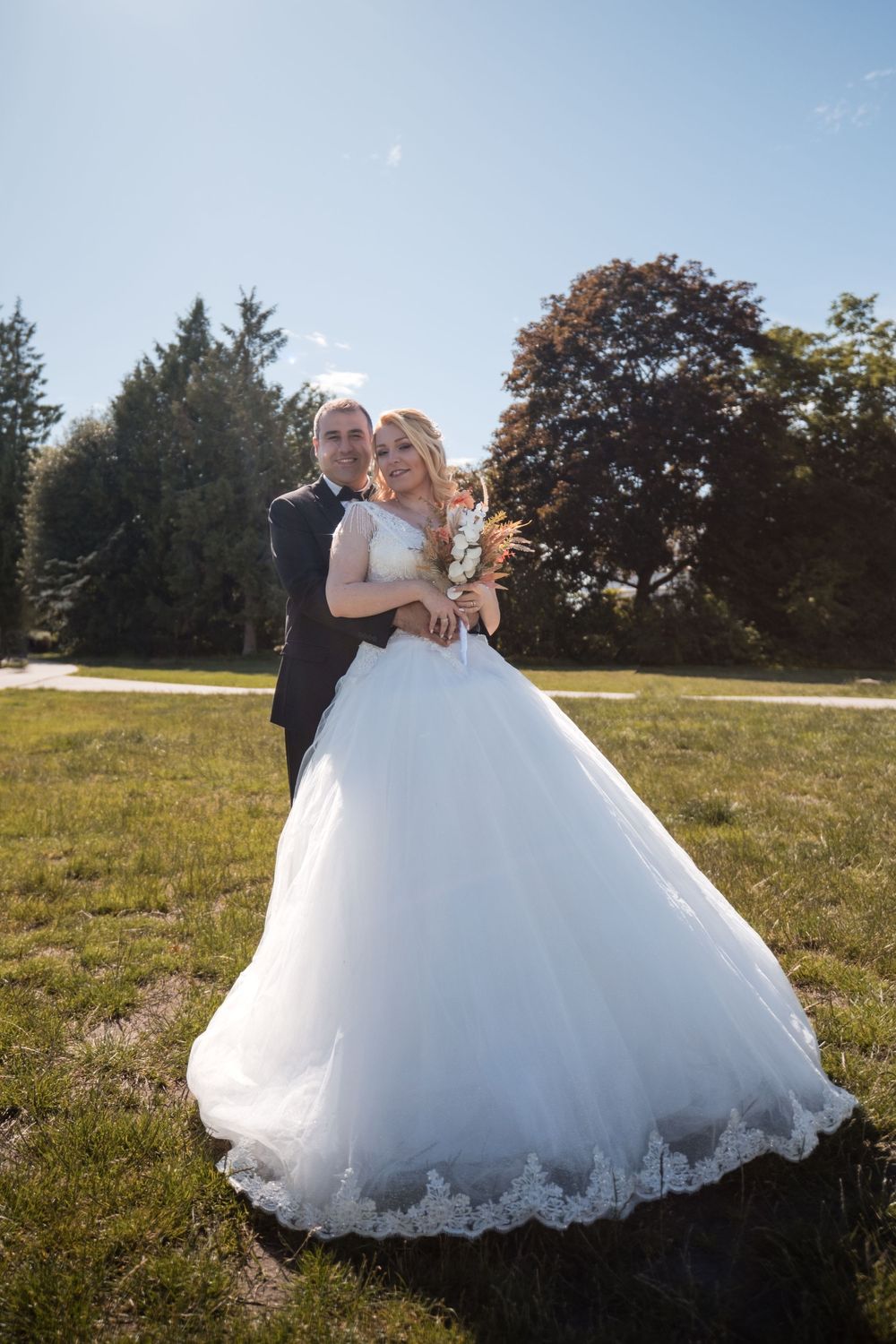 Newlywed couple embraces in a sunny meadow with autumn trees in the background.