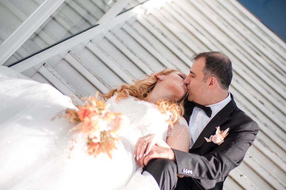 A couple in wedding attire share a romantic dip kiss in front of a white wooden building.
