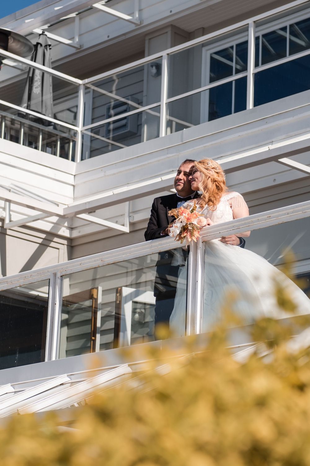 Wedding couple shares a romantic kiss on a balcony with yellow foliage in the foreground.