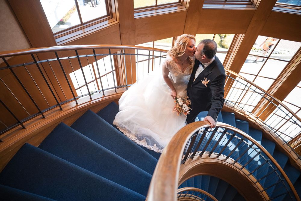 A couple in formal wedding attire dance together in a rustic wood-paneled venue with natural lighting from above.