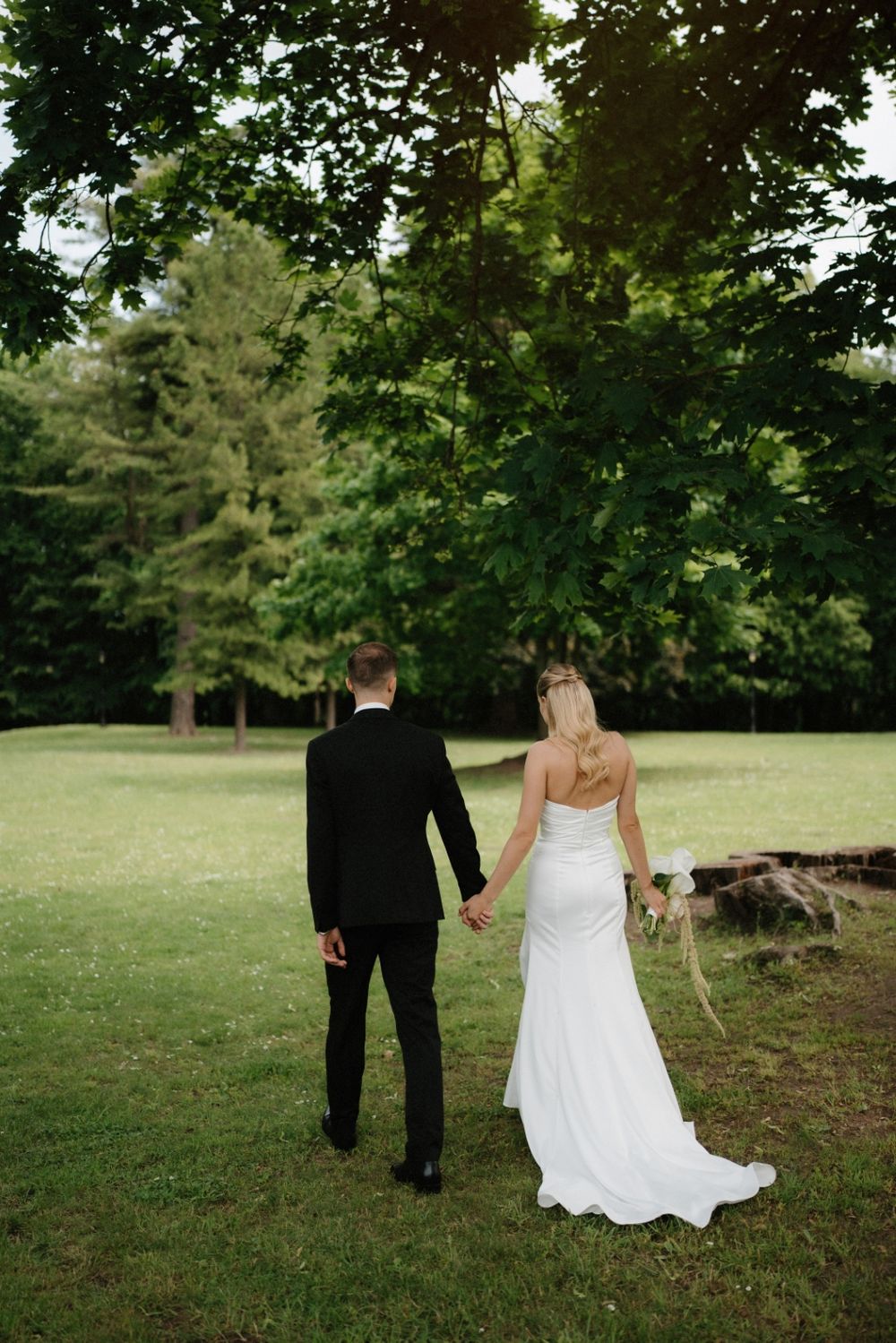 A couple in wedding attire walk together on a lush green lawn surrounded by trees.