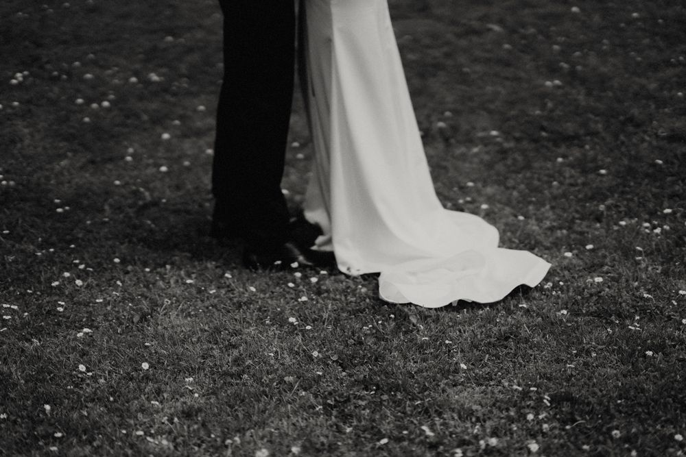 Black and white photo showing the flowing train of a wedding dress on grass.