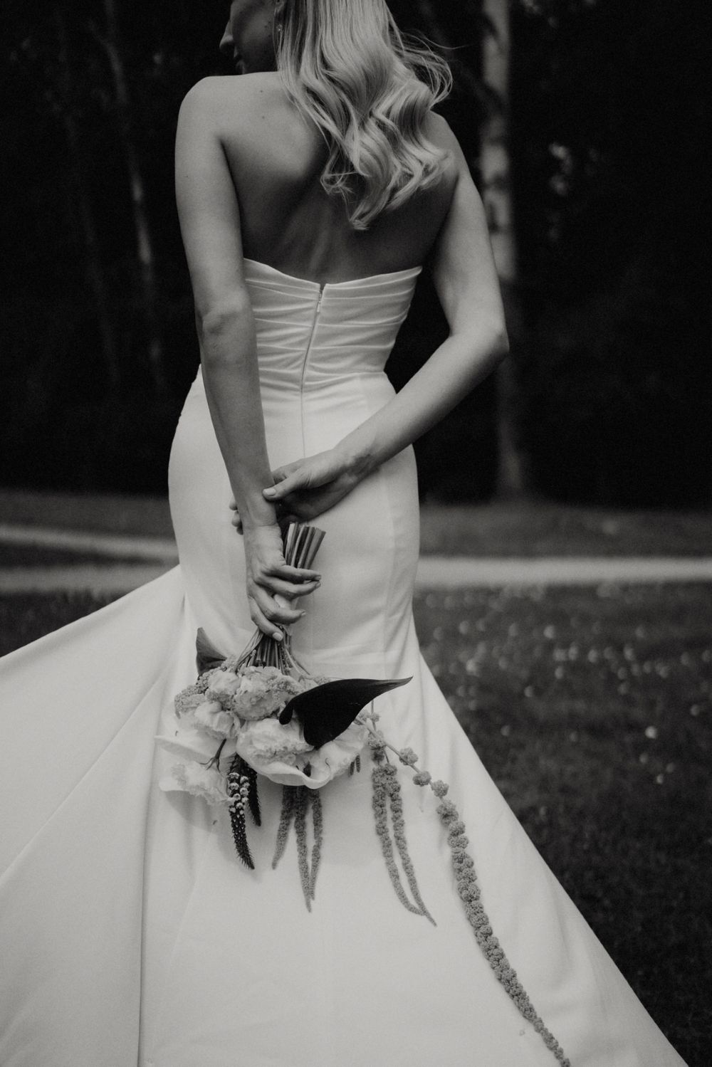 Black and white detail shot of a bride's dress and bouquet from behind.