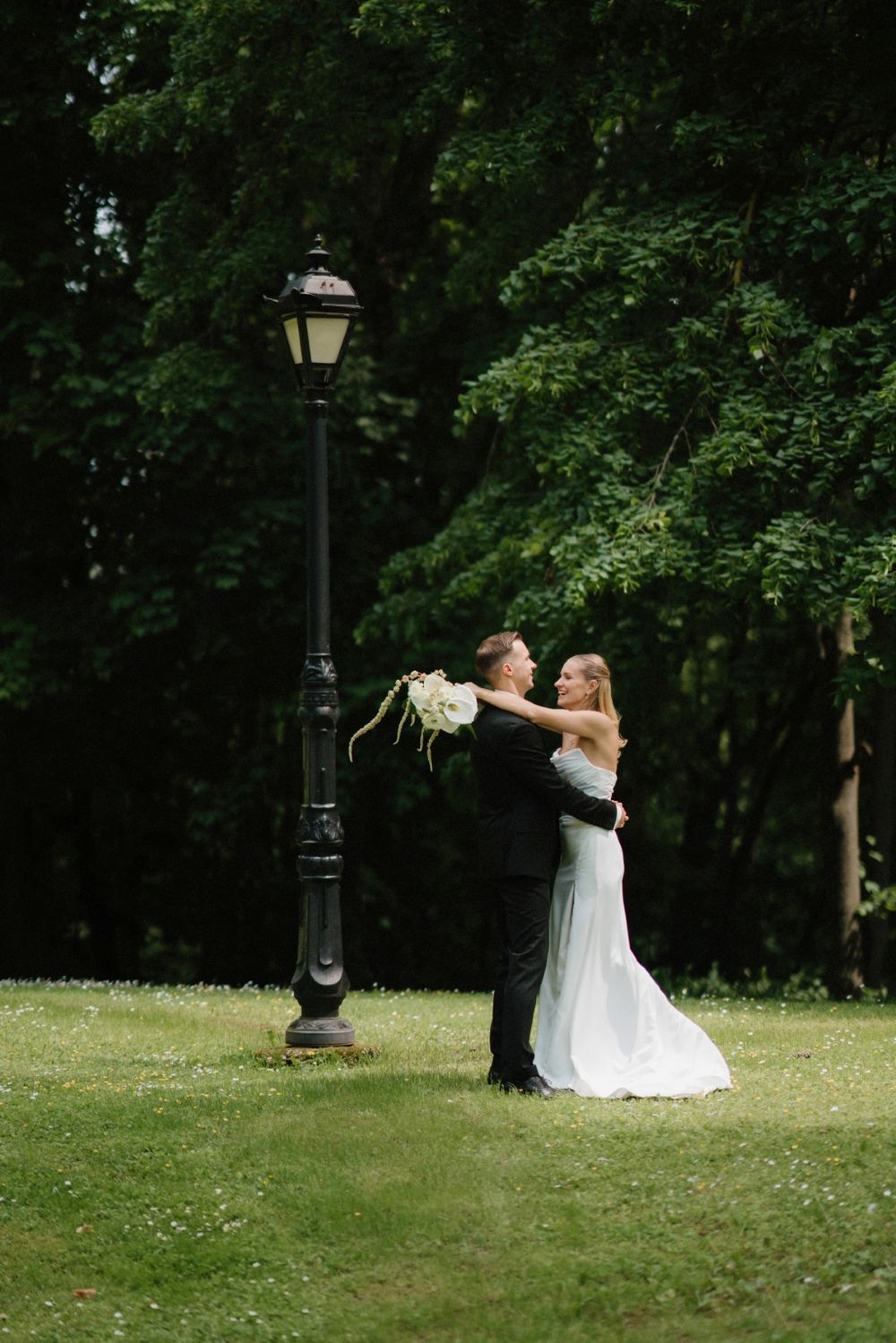 Wedding couple shares a playful moment on a grassy lawn with vintage lampposts.